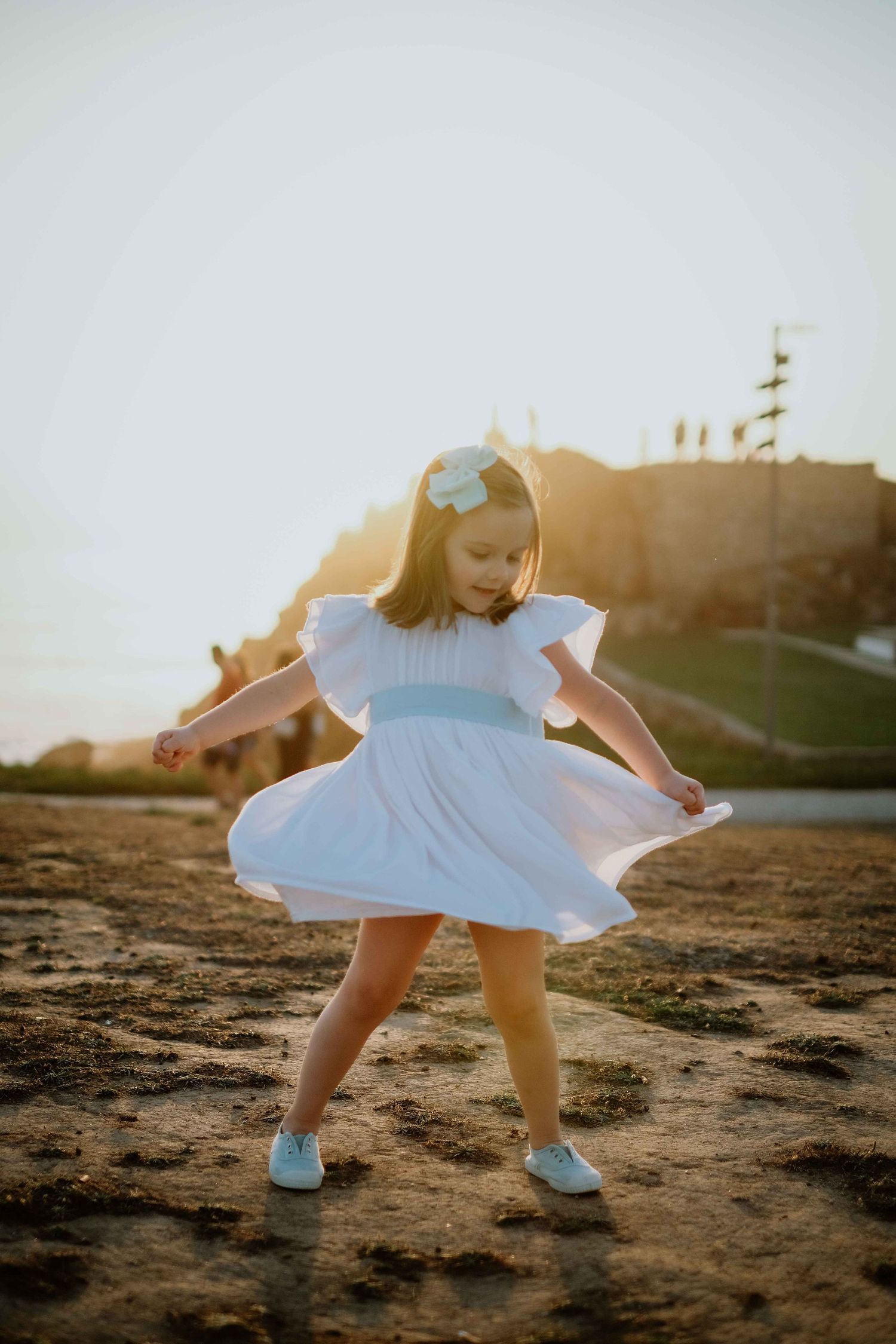 Child in white dress bending down to explore sandy ground at sunset.