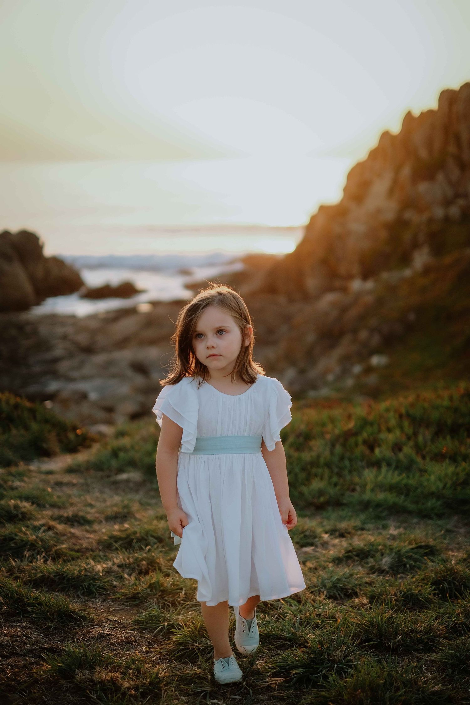 Little girl in white dress explores rocky coastal area at sunset.
