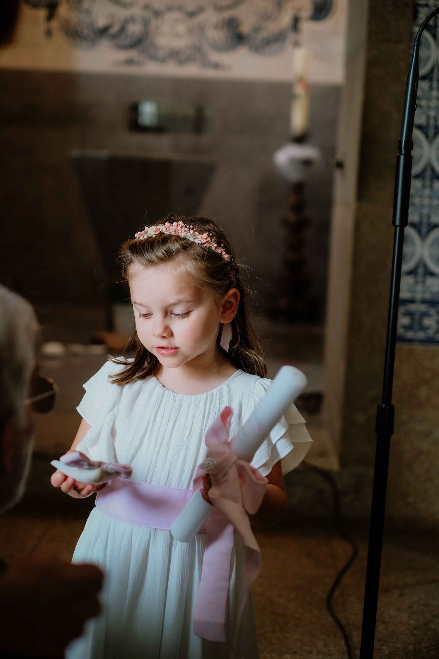 Young child in white dress with pink sash stands in ornate interior setting.