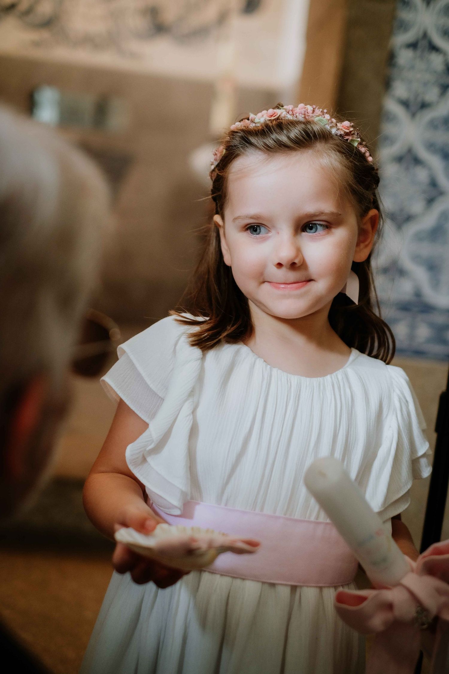 Child wearing white dress with pink sash poses in elegant interior setting.