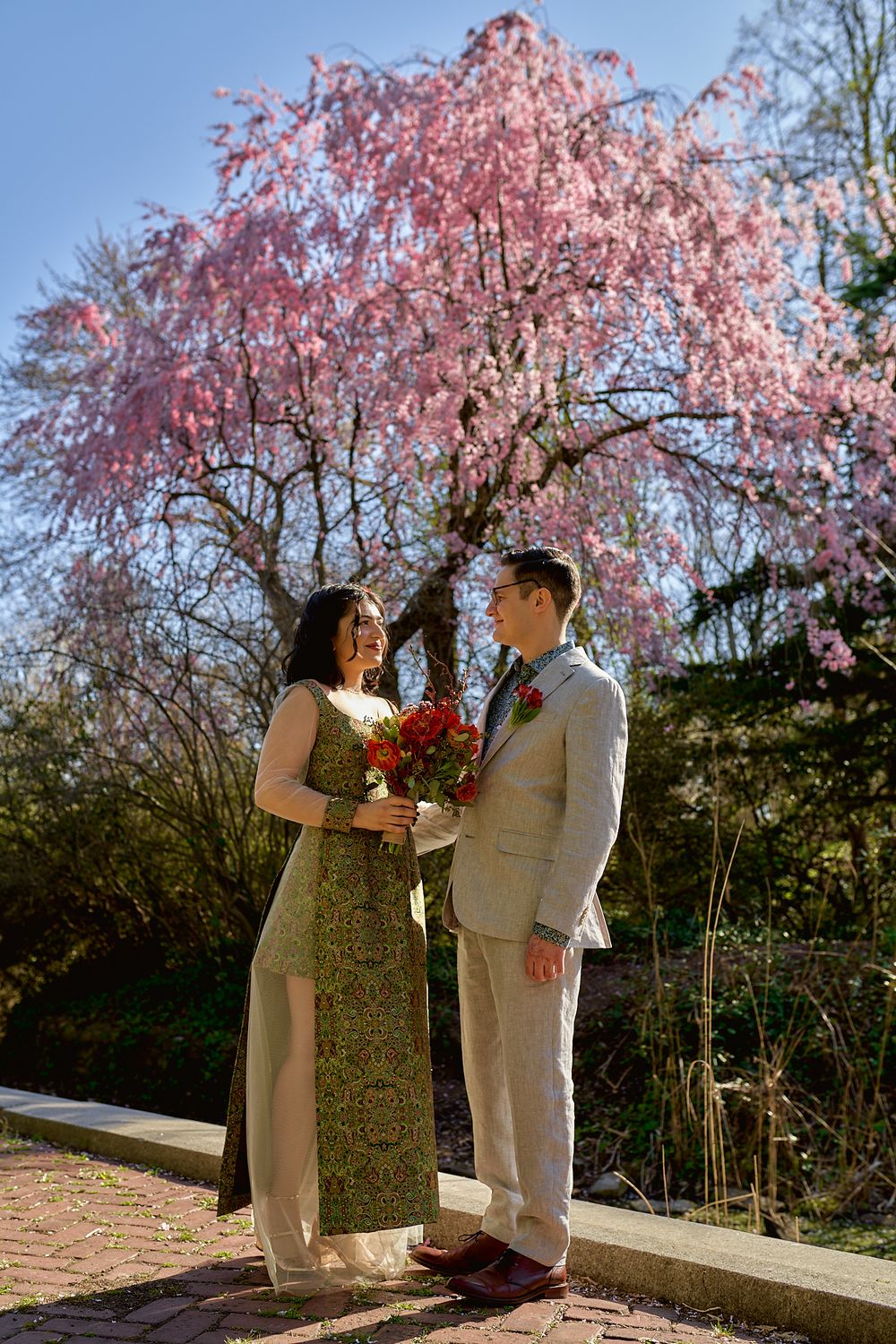 A couple poses beneath a blooming cherry blossom tree holding a vibrant bouquet.