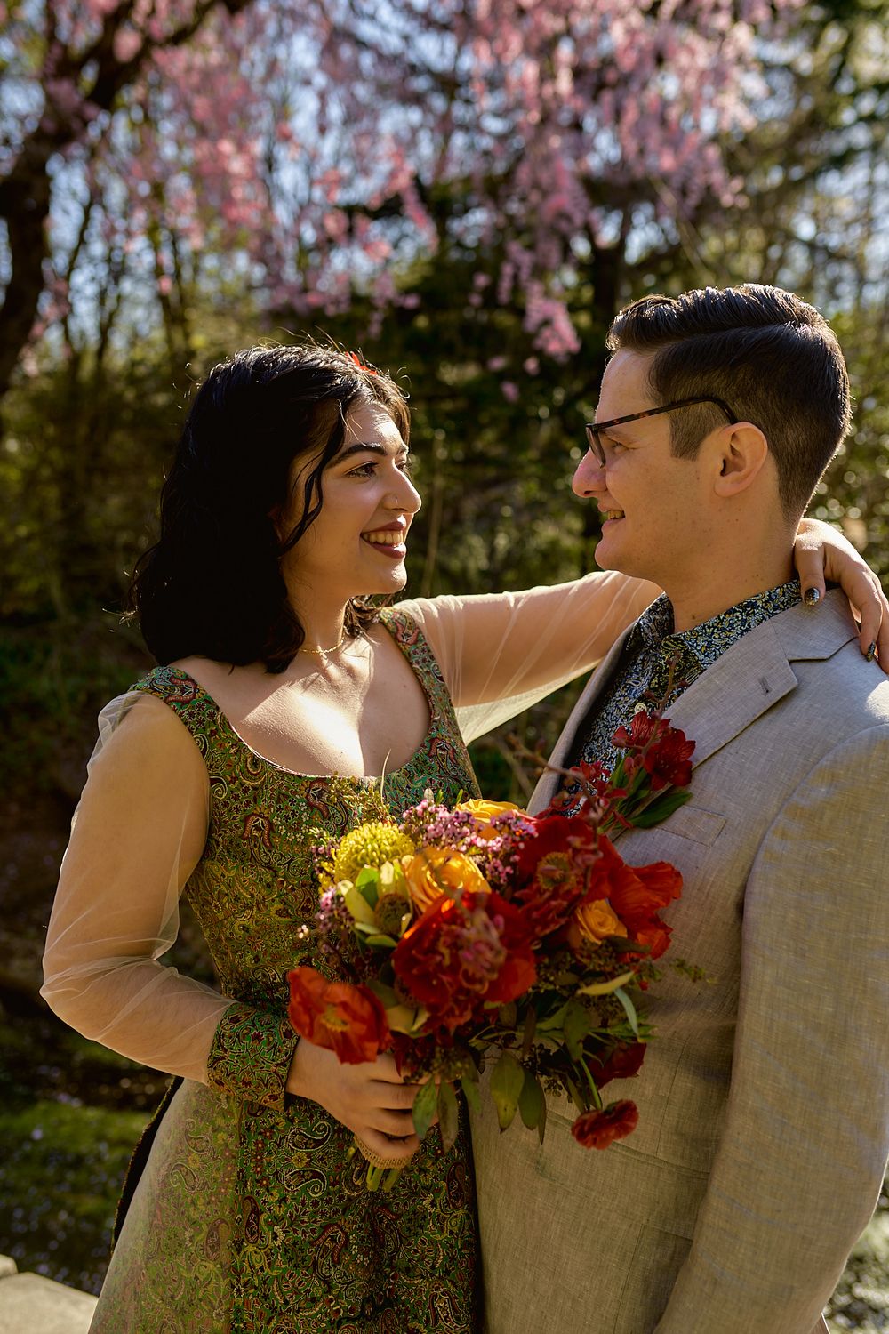 A couple shares an intimate moment under cherry blossoms with a colorful floral bouquet.