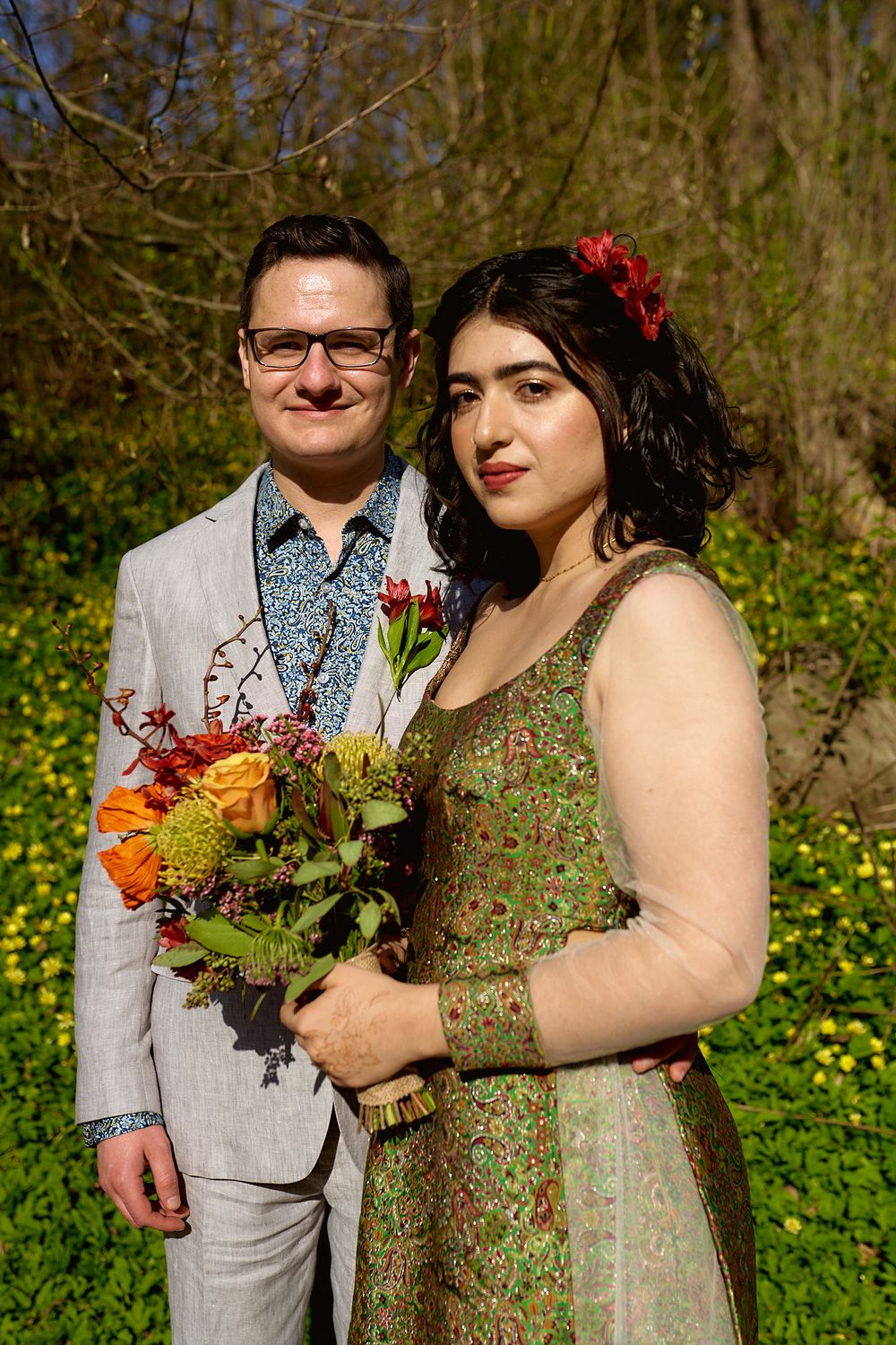A couple poses in formal attire with a bright autumn-colored bouquet in a garden setting.