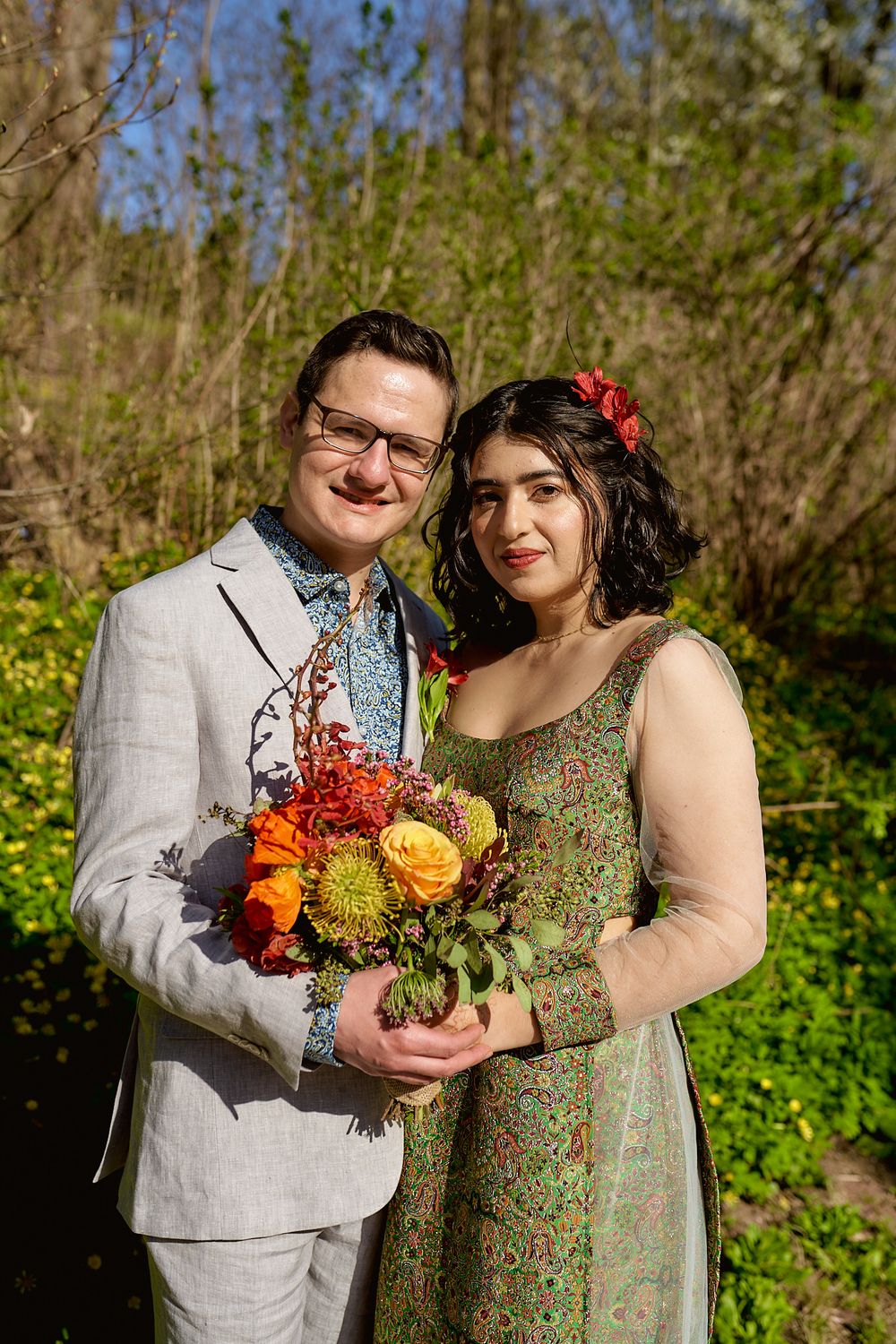 A couple smiles together in a garden while holding a vibrant bouquet of orange and red flowers.