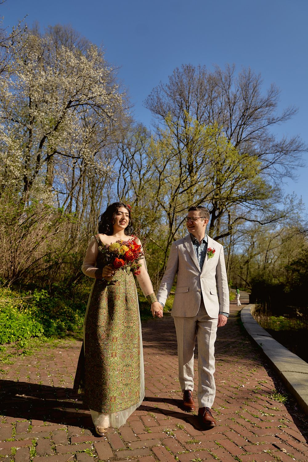 A couple strolls together on a brick pathway under budding spring trees.