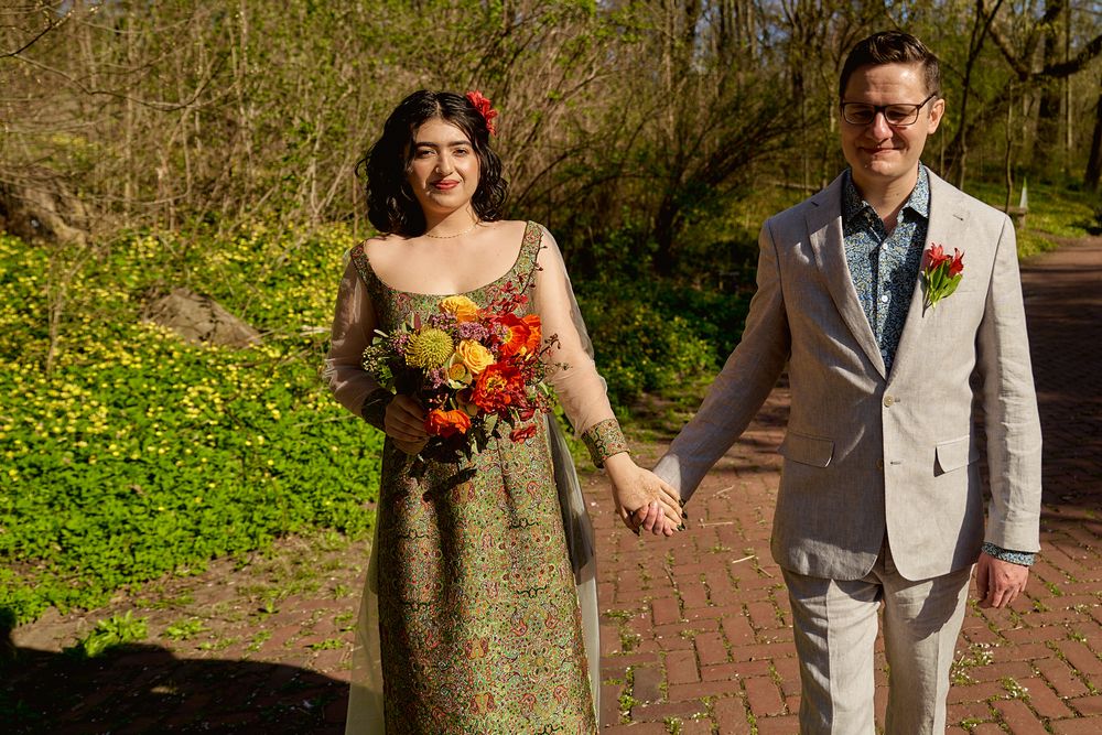 A couple holds hands outdoors in spring, wearing formal attire with a colorful bouquet against a natural backdrop.
