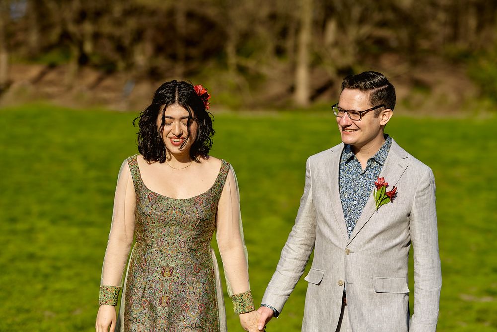 A couple walks hand in hand across a sunny green lawn in formal attire.
