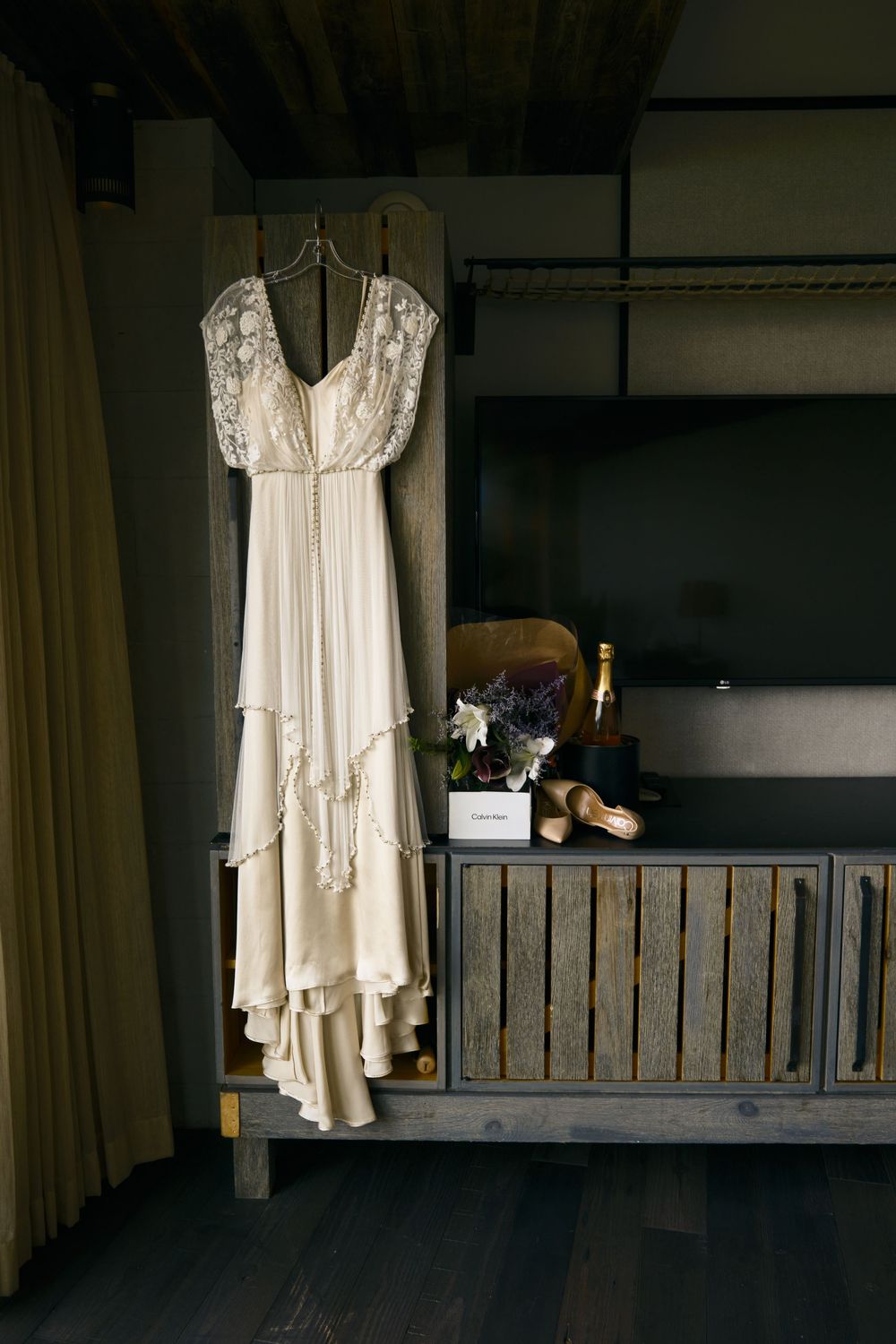 A vintage-style lace wedding dress hangs elegantly against a rustic wooden wall with flowers and decor.