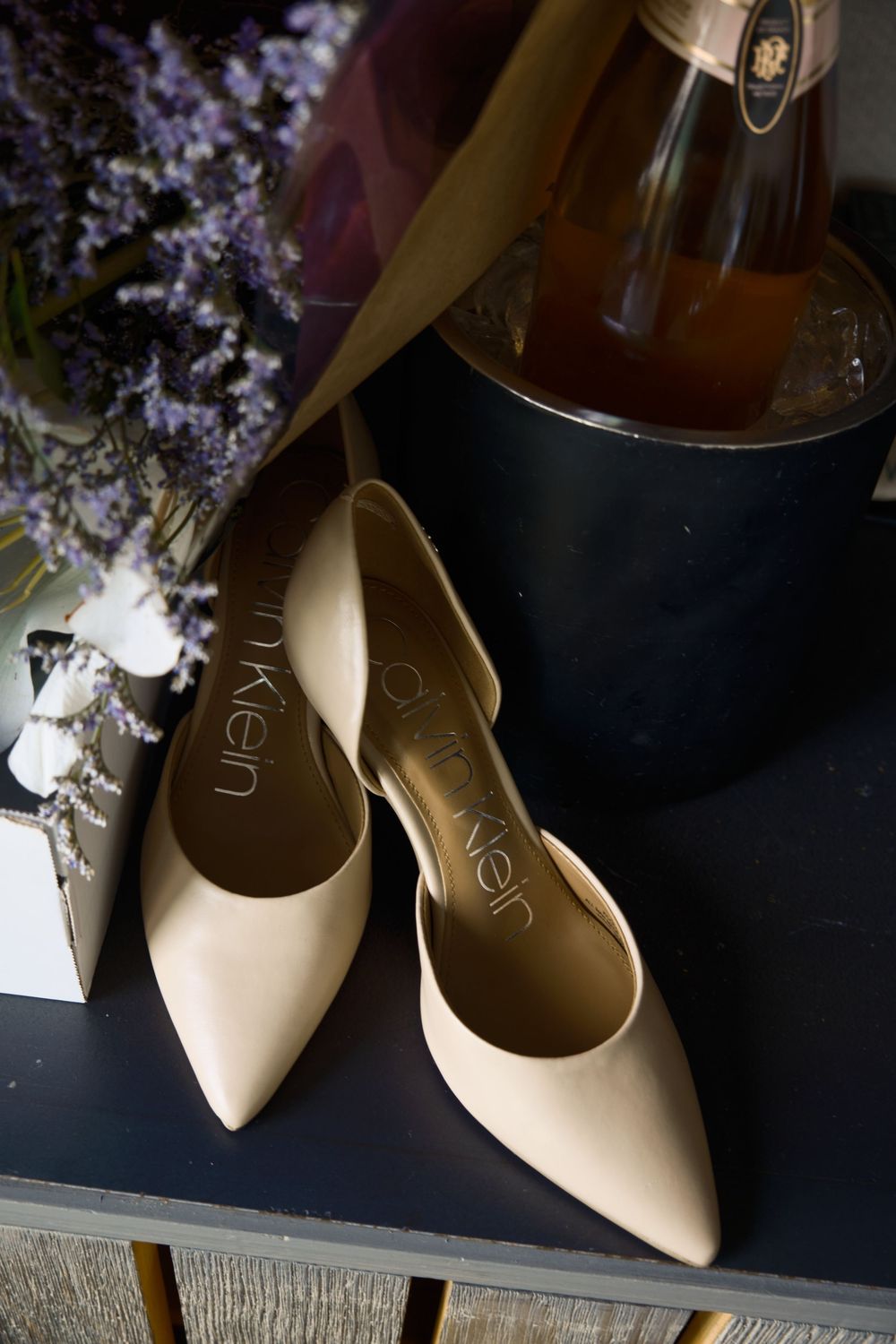 Cream colored bridal shoes displayed with lavender flowers and champagne bottle.