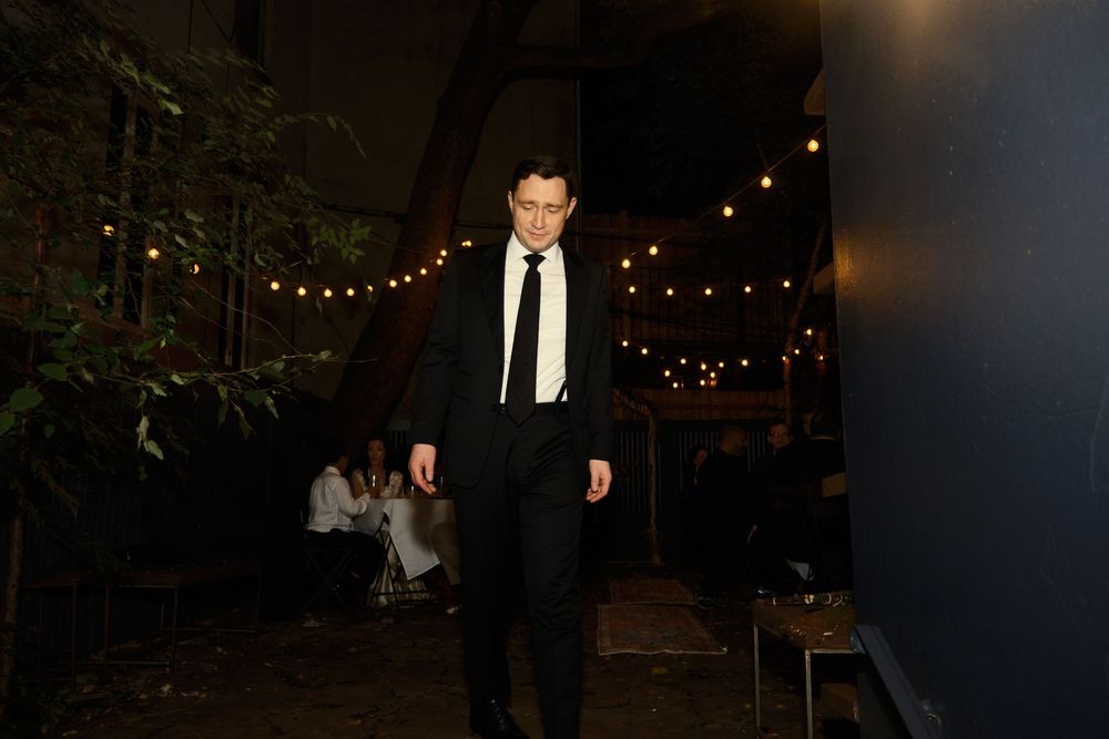 A person in formal attire stands alone under string lights at an outdoor evening event at Mayson May