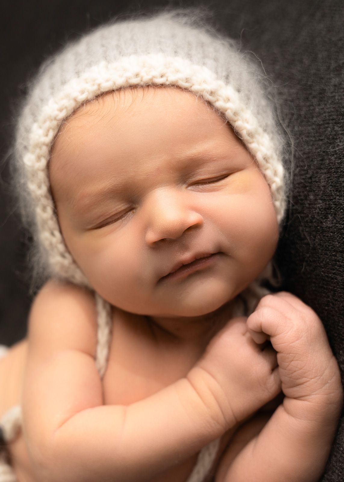 A sleeping baby wearing a white knit hat rests peacefully with their thumb in their mouth.
