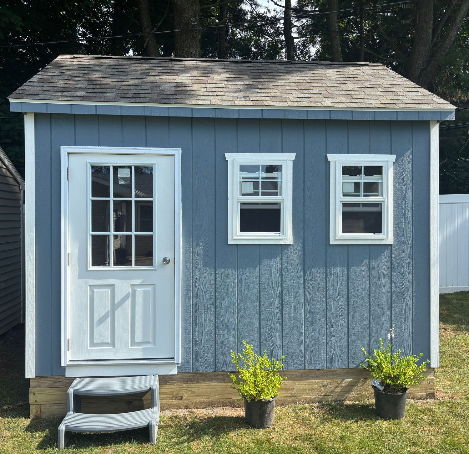 Small blue-gray wooden shed with white trim, door, and two windows surrounded by green landscaping.