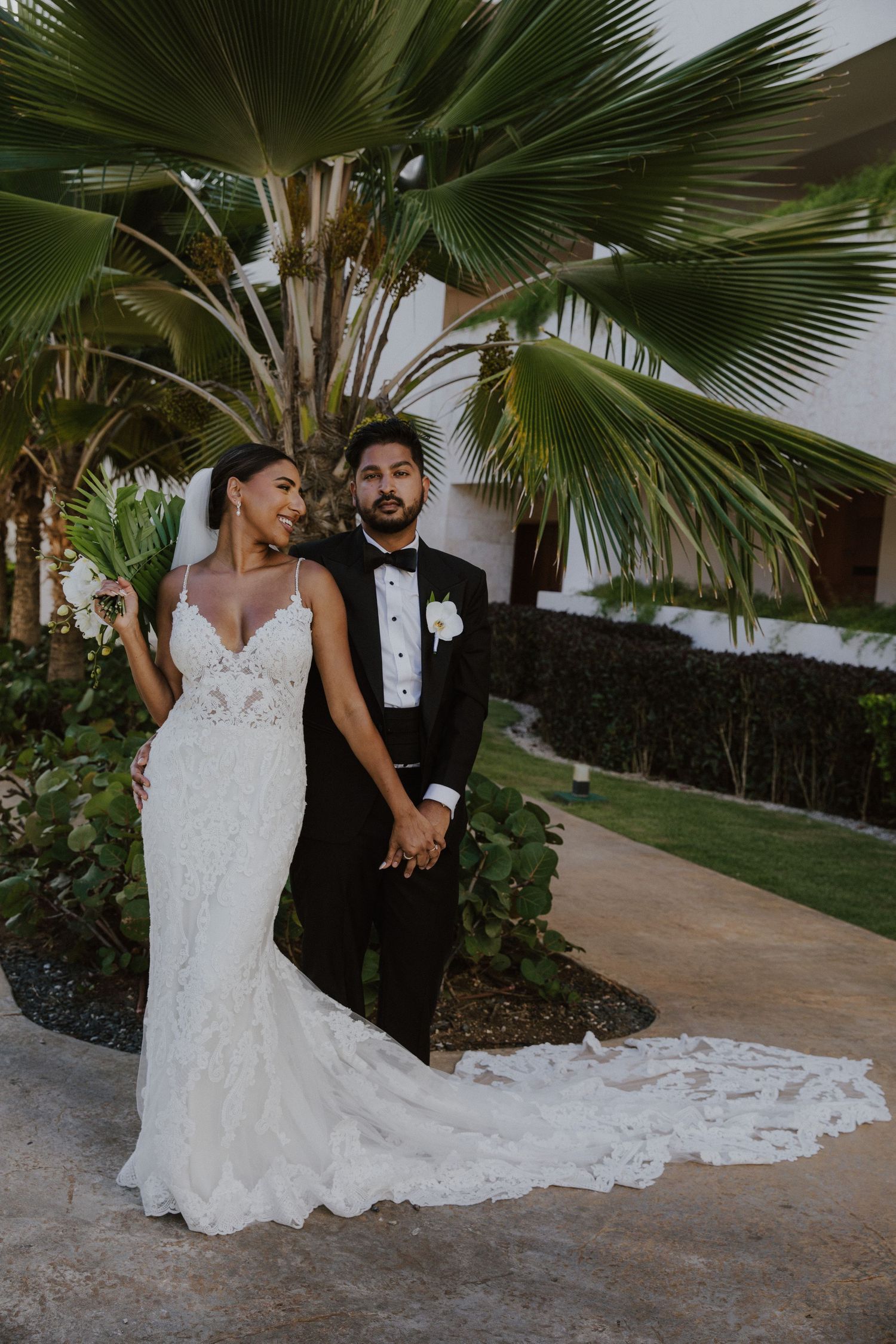 Newlyweds holding hands under palm trees, bride in detailed lace gown with long train.