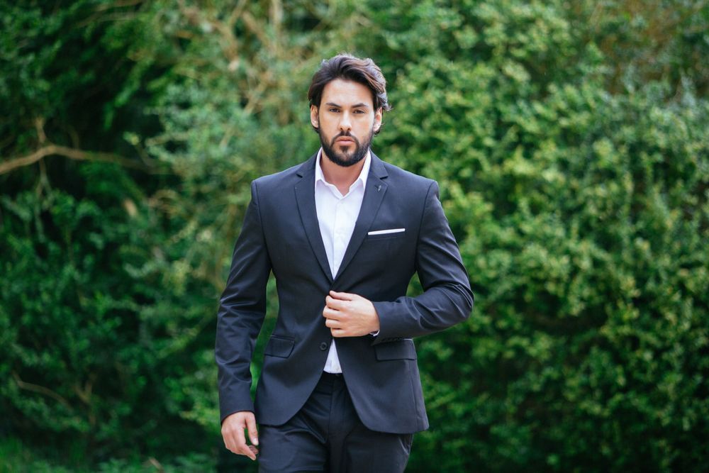 A man in a navy business suit and white shirt poses confidently against a green outdoor garden background.