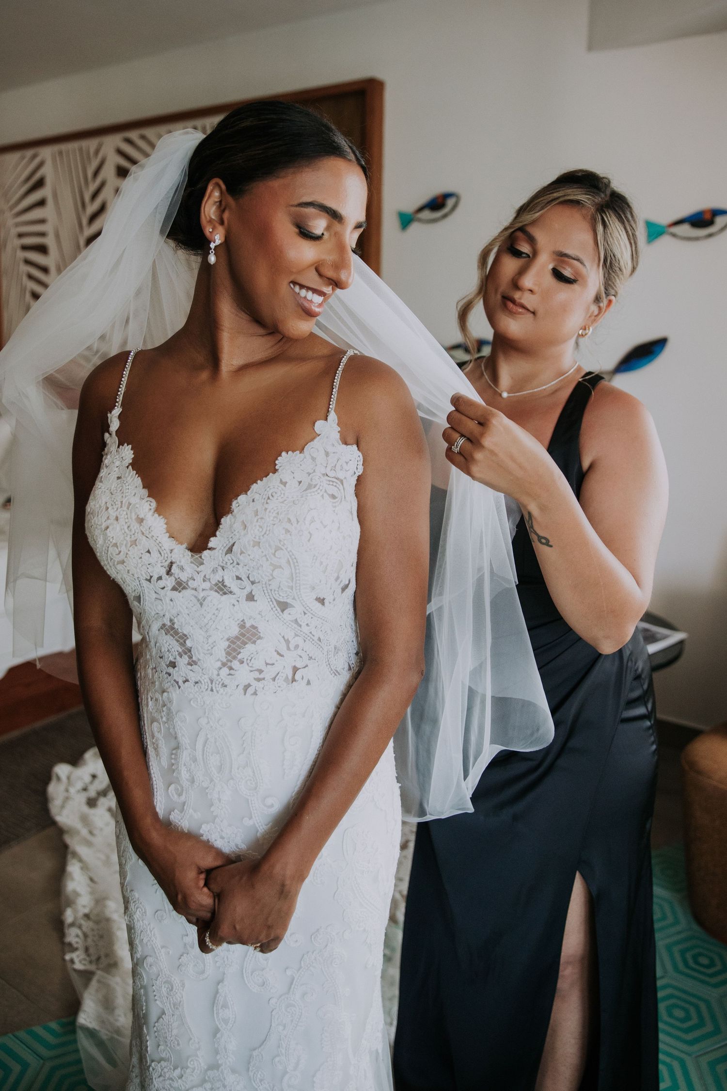 Bride in intricate lace wedding dress smiles while bridesmaid adjusts her veil.