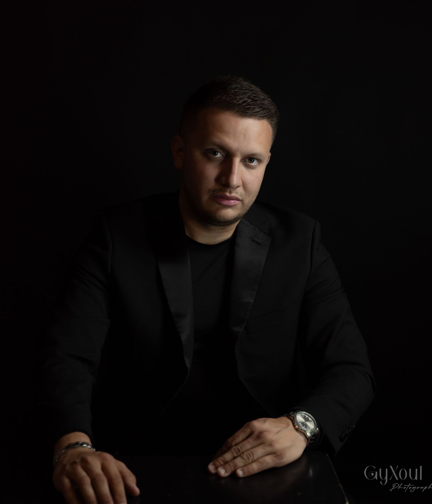 Professional dark studio portrait showing a person in a black suit jacket against a dramatic black background.