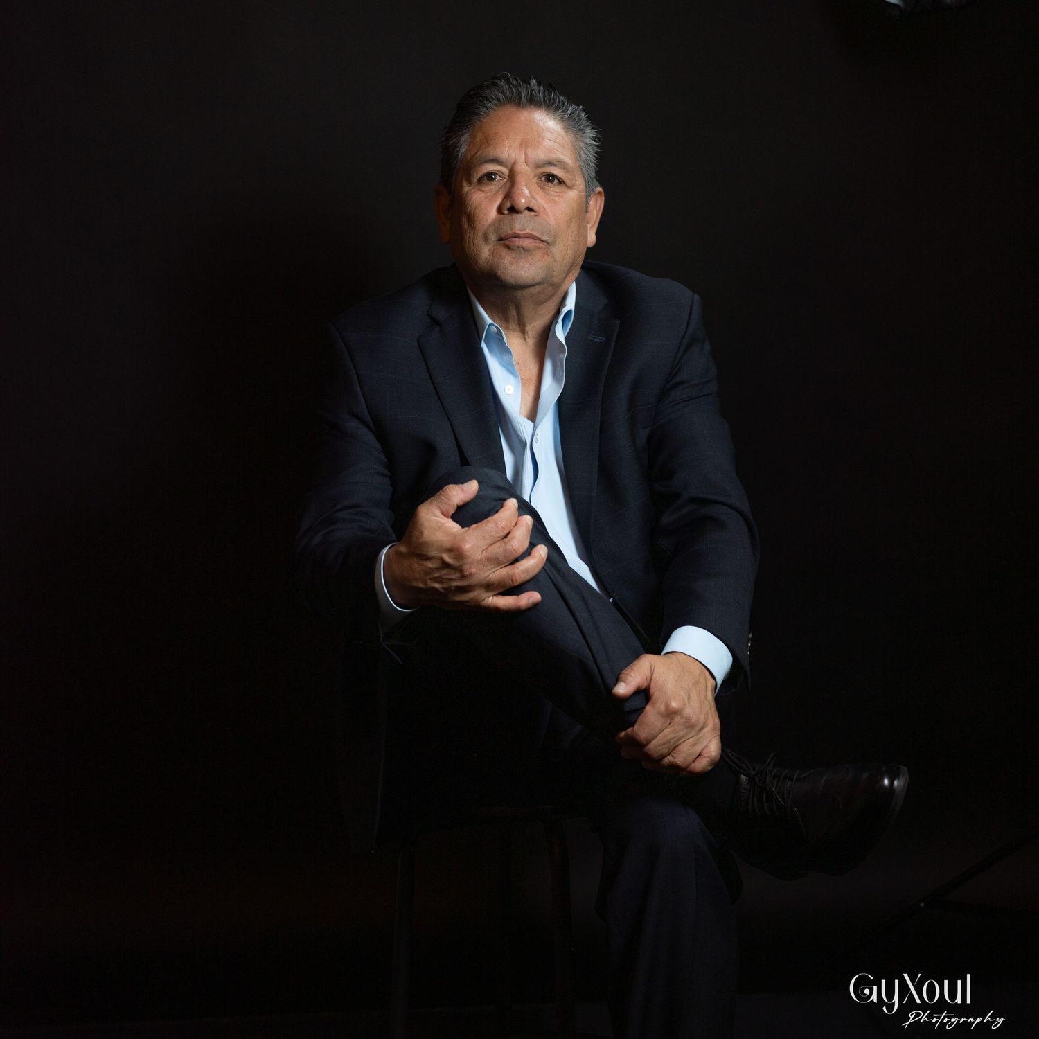 Professional studio portrait of someone in a dark suit and white shirt seated against a black background in dramatic lighting.