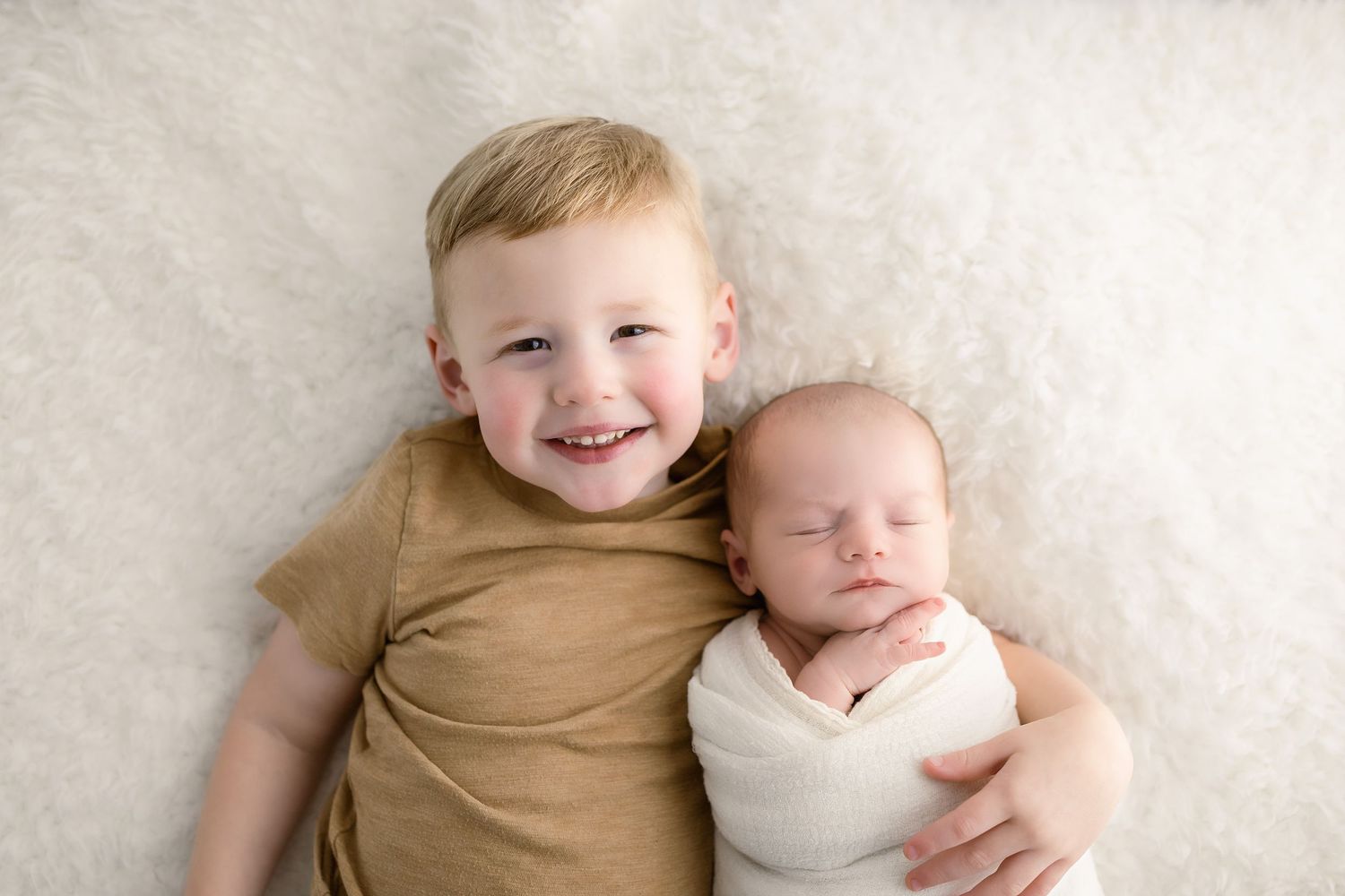 Two young children lying together on a beige backdrop, one wearing tan and one in white.