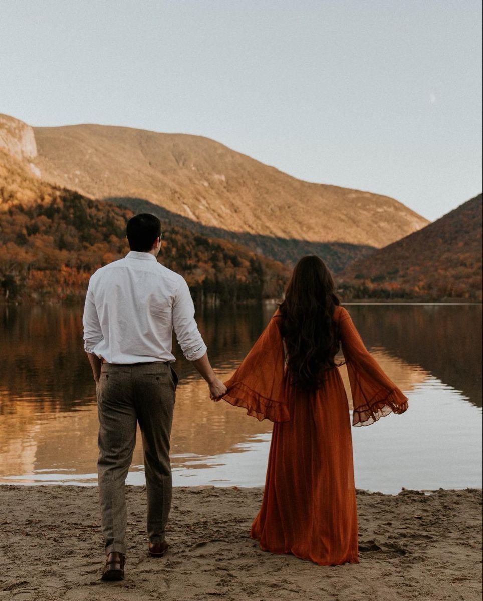 A couple in autumn attire sits together by a tranquil mountain lake at sunset, taking in the serene landscape.