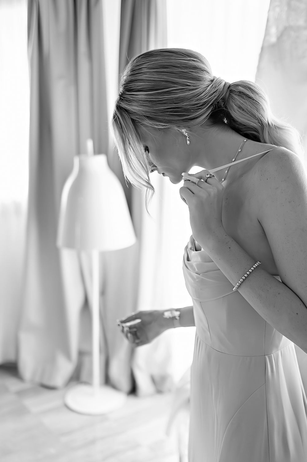 Black and white photo of a person adjusting a white dress while standing near a floor lamp in an elegant room.