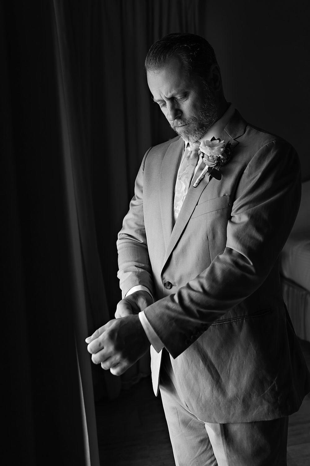 Dramatic black and white portrait of a groom adjusting his cuff in window light.