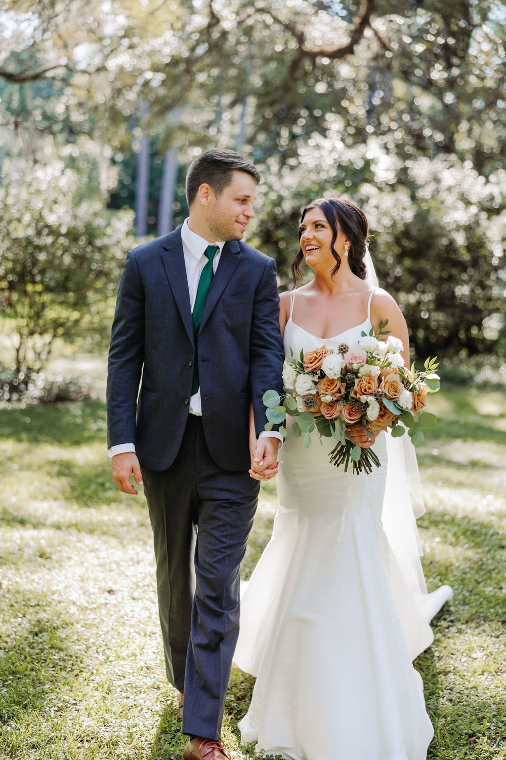 A couple in wedding attire, with a navy suit and white dress, walk and laugh together in a sunlit garden setting.