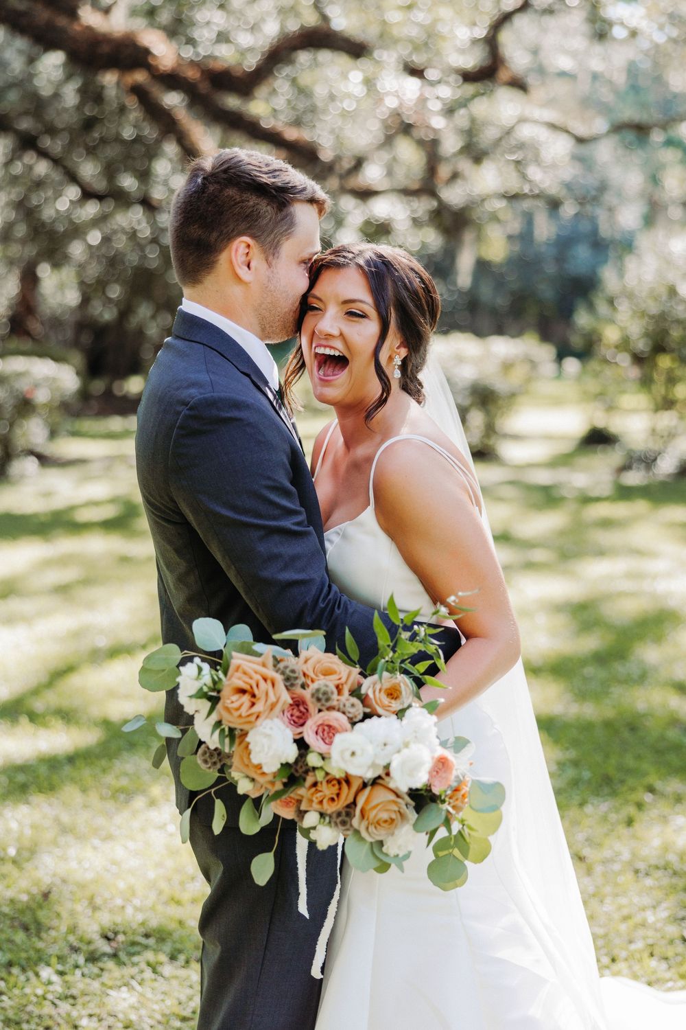 Joyful wedding couple shares a sweet moment outdoors with a beautiful peach and pink floral bouquet.