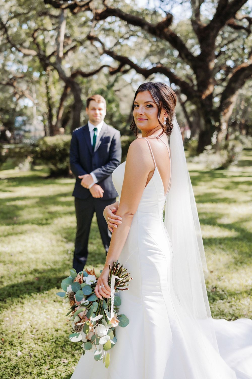 A couple poses under majestic oak trees at their outdoor wedding, with the bride holding a eucalyptus bouquet.