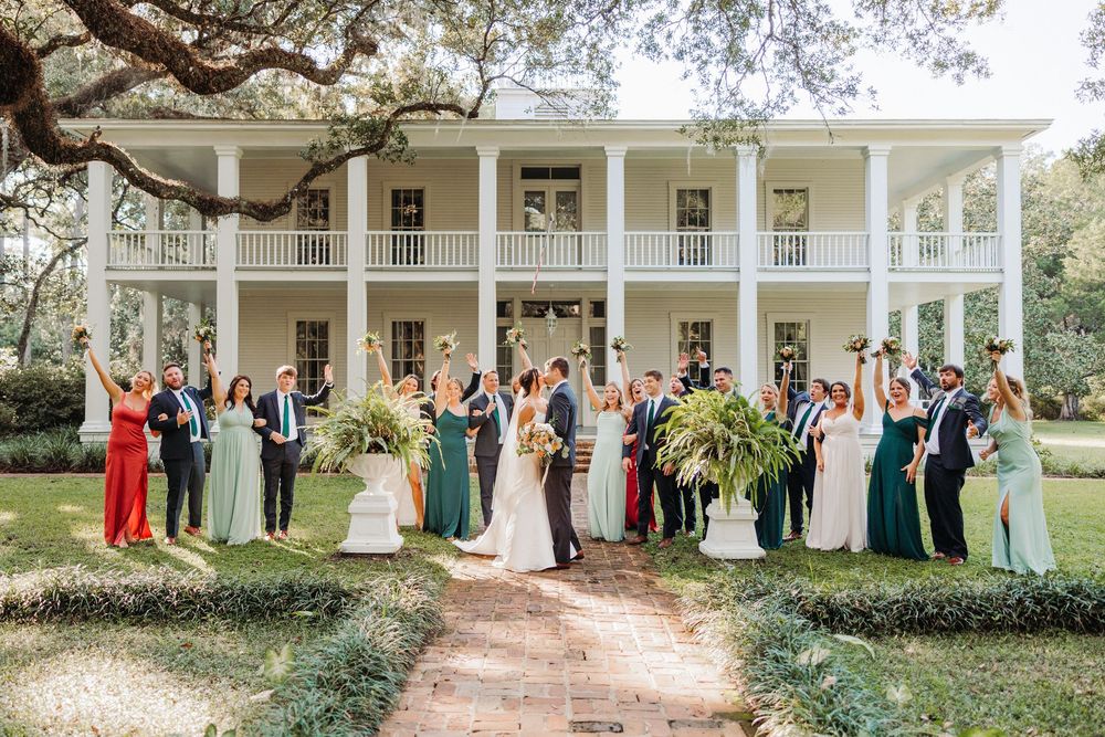 Wedding party poses in front of a grand white antebellum style mansion with tall columns and wraparound porches.