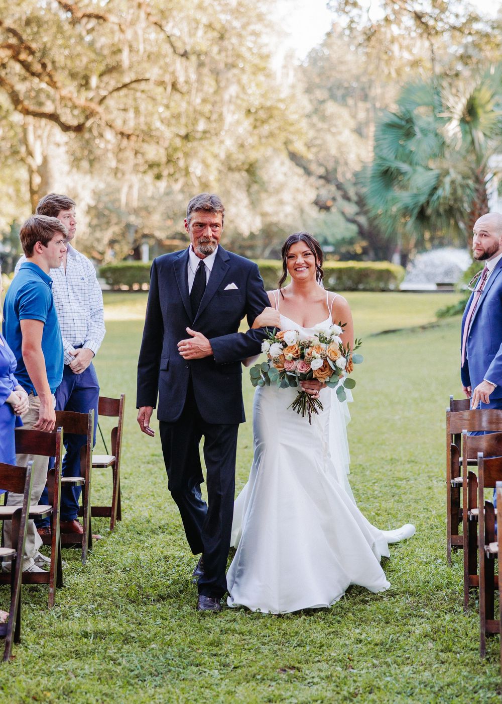 Wedding guests share joyful moments amid wooden chairs set up on a grassy lawn during an outdoor ceremony.