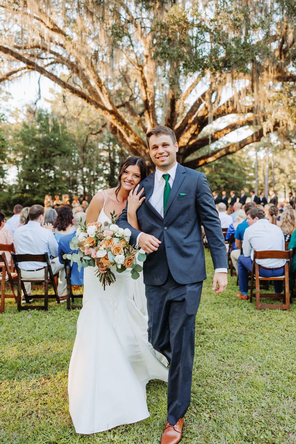 Wedding couple shares candid moments while walking and laughing together under moss-covered trees at an outdoor reception.