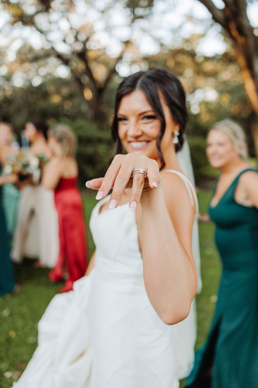 A bride in a white wedding dress holds up her hand to show off her ring during outdoor festivities.