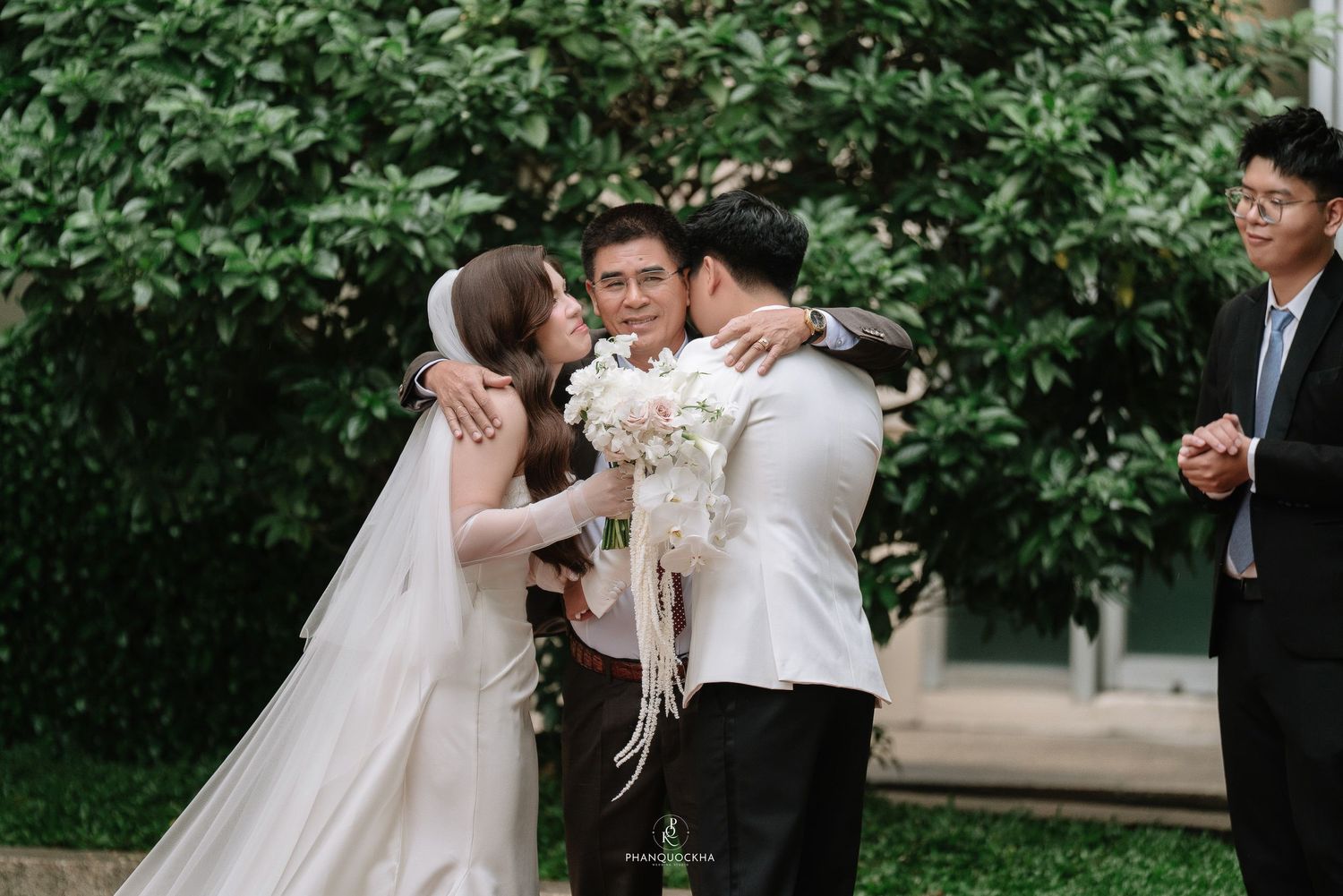 Bride in white gown kisses elder guest on cheek while groom stands beside her at outdoor wedding.