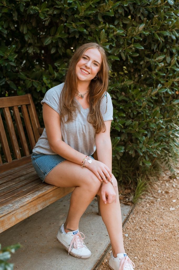 A person in a gray t-shirt and denim shorts sits on a wooden bench in front of green leafy bushes.