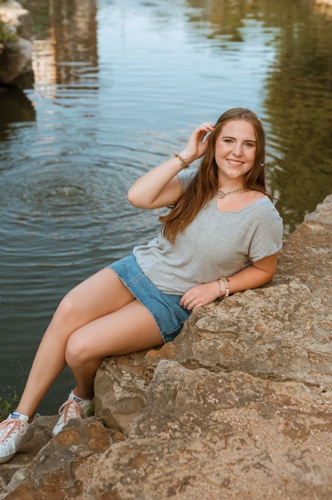 A smiling person in a gray t-shirt and jean shorts sits on rocks by a calm pond on a sunny afternoon.