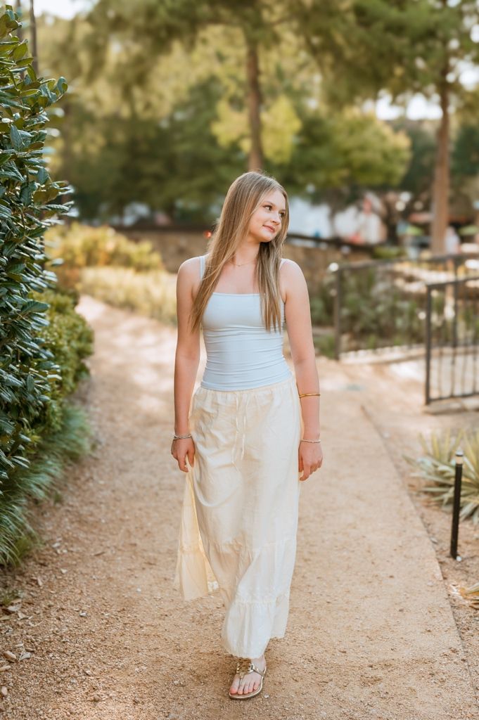 Person in a light blue tank top and flowing cream skirt walks along a scenic garden path on a sunny afternoon.
