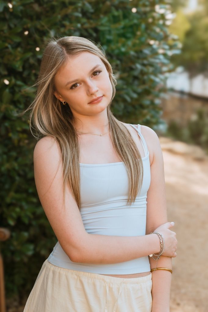 Person in a light blue sleeveless top poses against green foliage in warm natural lighting.