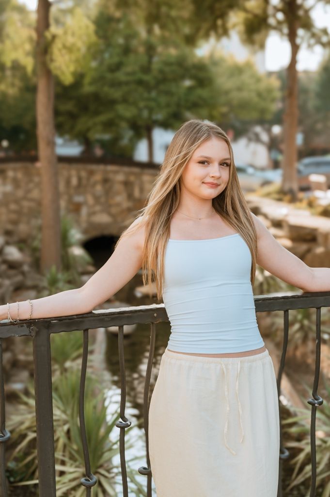 Young woman in light blue top and cream skirt poses gracefully against wooden railing at outdoor location during sunset.