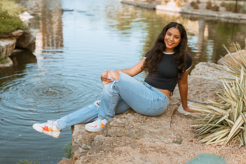 Woman wearing blue jeans and black top posing by water's edge with dangling legs during sunset photoshoot.