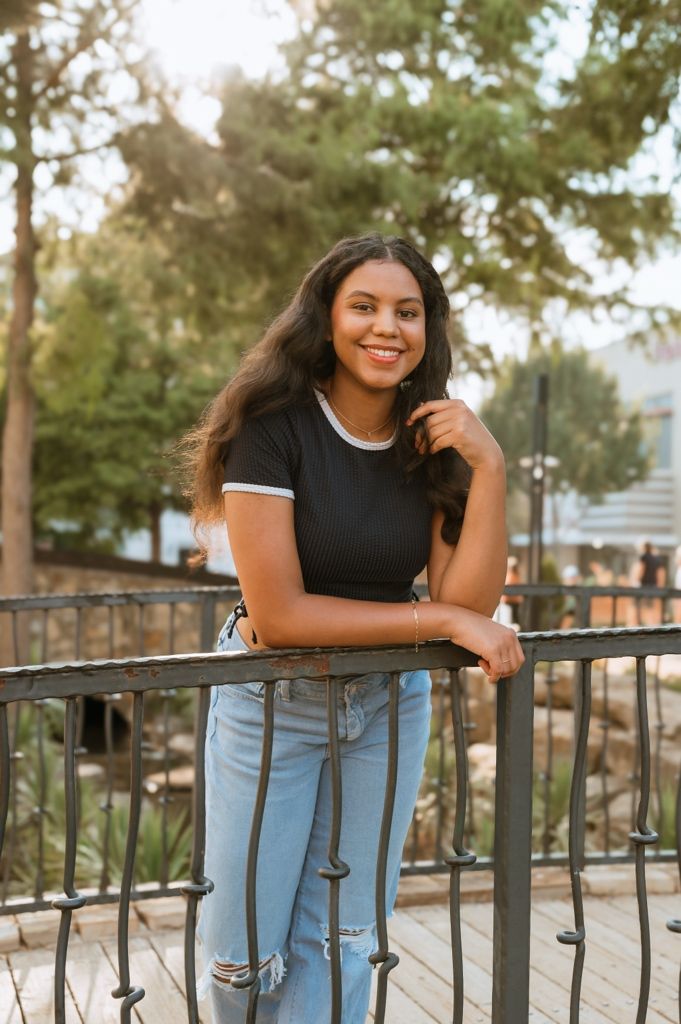 Young woman in black t-shirt and ripped jeans poses smiling on outdoor balcony during golden hour.