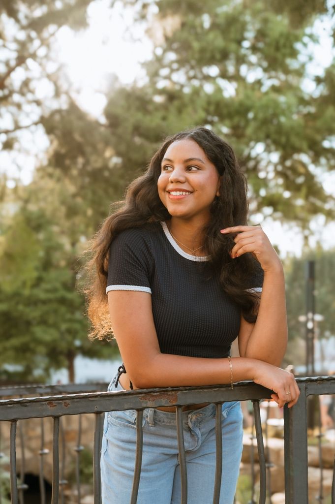 A person in a black ribbed t-shirt with white trim leans on a railing outdoors, smiling warmly in golden sunlight.