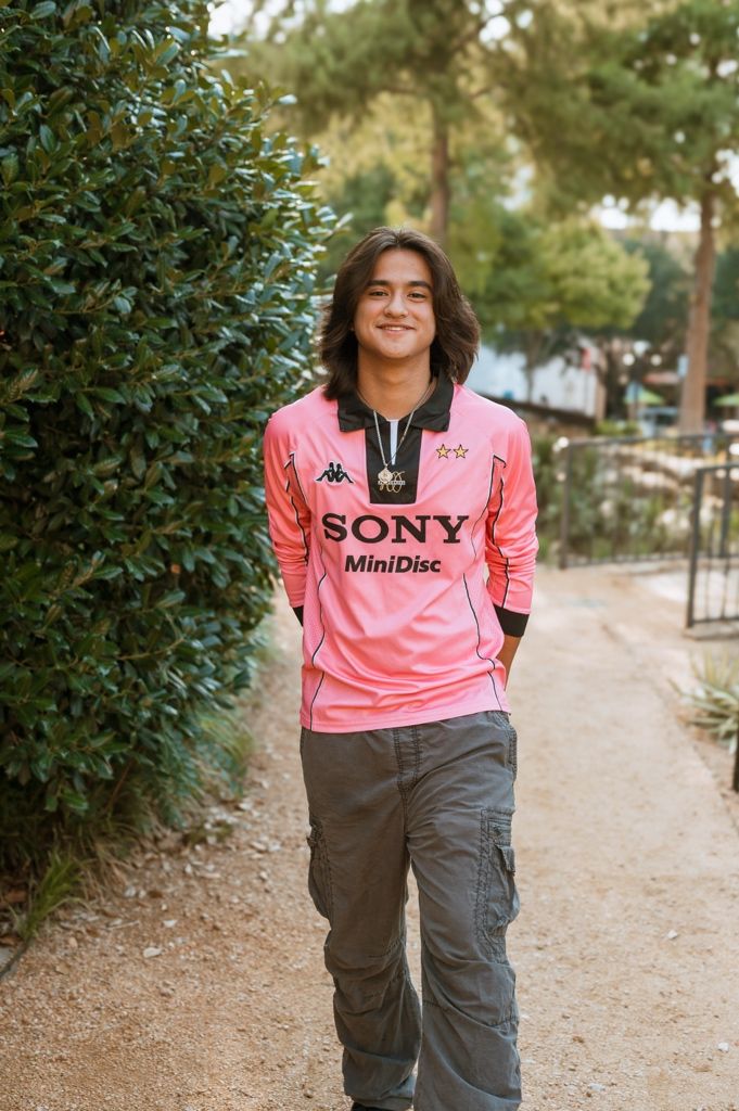 Someone wearing a pink Sony MiniDisc jersey stands casually on a path near green foliage on a sunny day.
