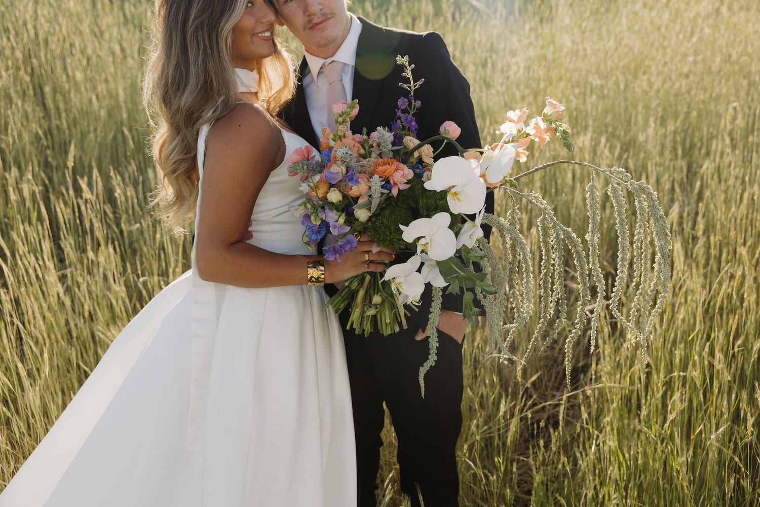Wedding couple embracing in golden wheat field, bride holding colorful bridal bouquet.