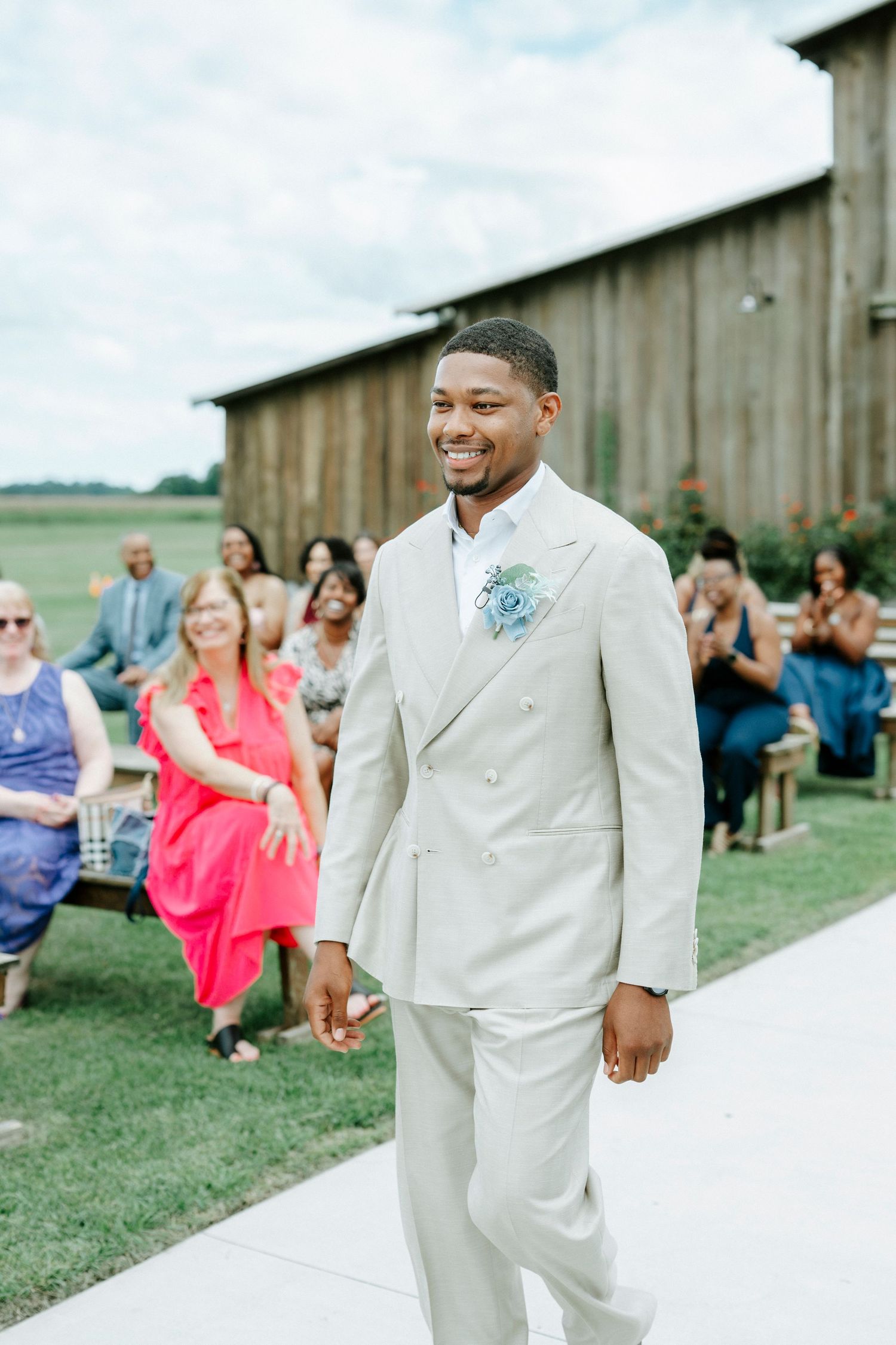 Groom in light colored suit stands before wedding guests at outdoor barn venue.