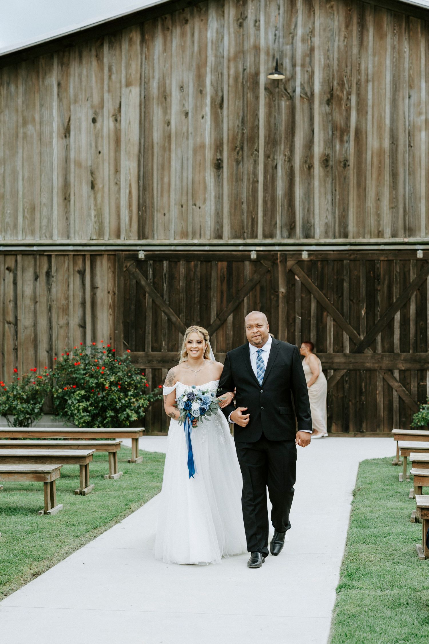 Wedding party member escorts bride down aisle with rustic barn backdrop.