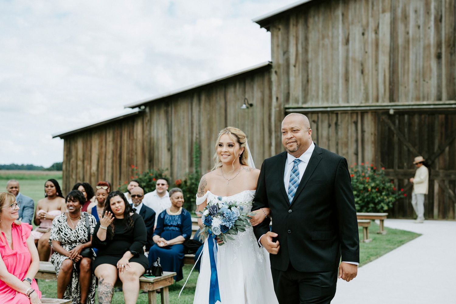 Outdoor wedding ceremony with rustic barn backdrop and guests seated on wooden benches.