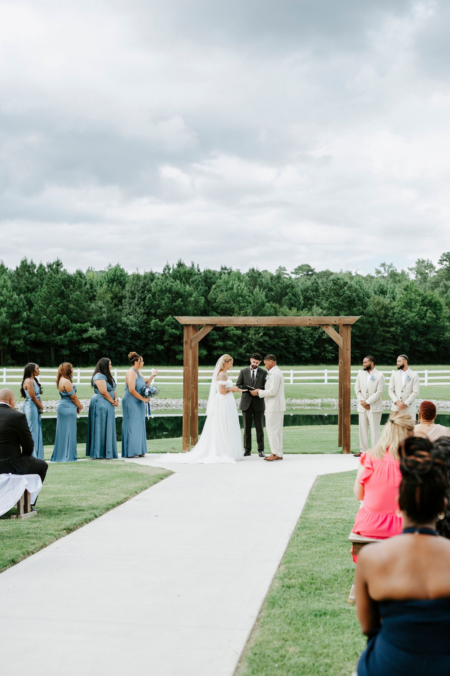 Wedding ceremony under wooden arbor with bridemaids in blue and scenic countryside view.