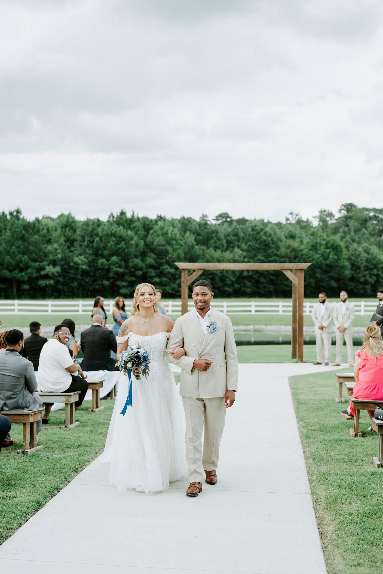 Newlyweds walk down white aisle runner at outdoor countryside wedding venue.