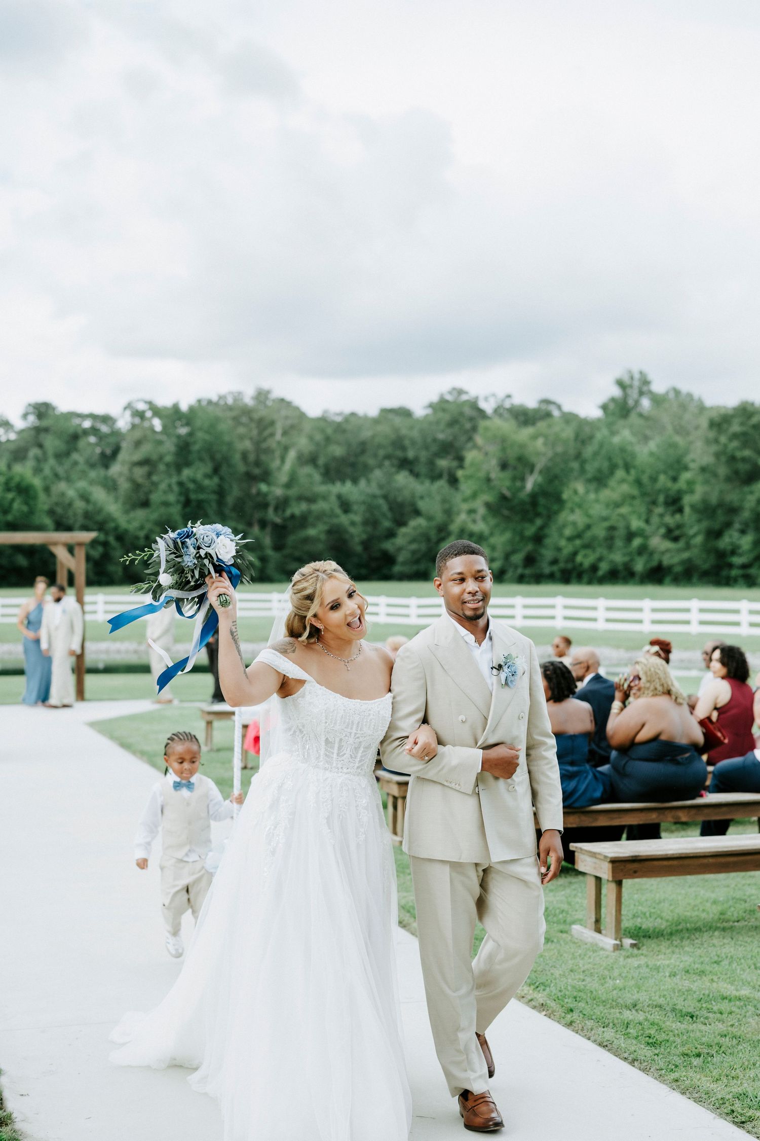 Happy couple walks down aisle at outdoor wedding with guests on rustic wooden benches.