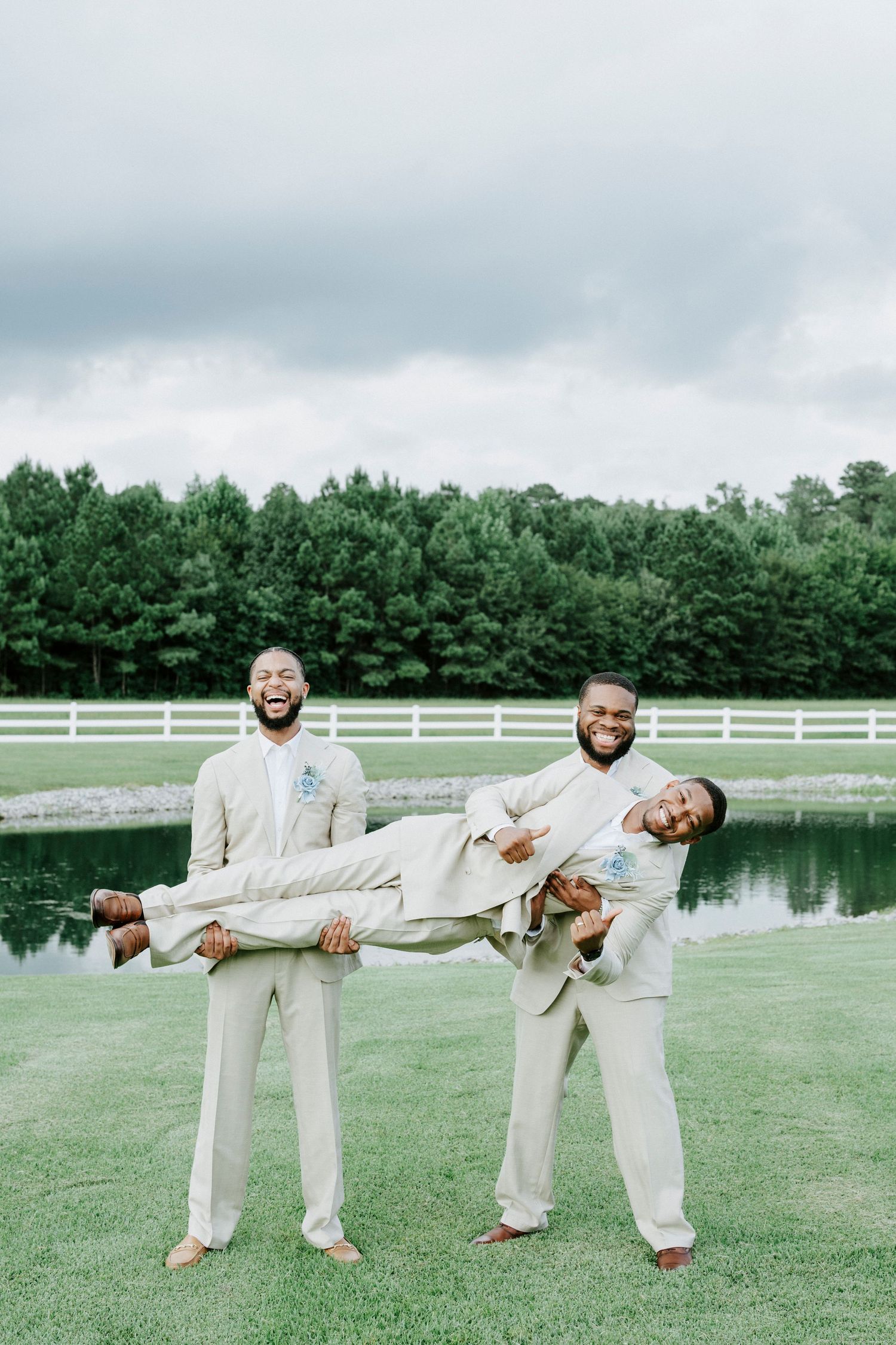Groomsmen in beige suits pose playfully on a green lawn with a pond and white fence backdrop.